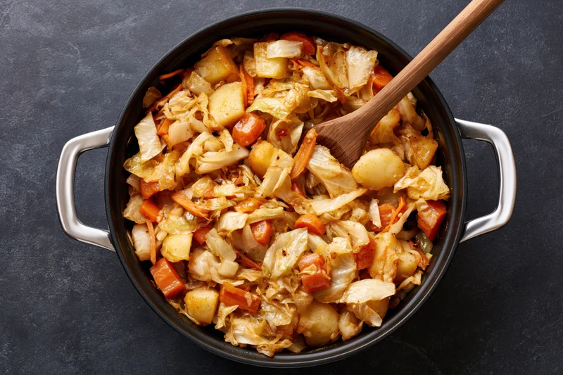 Sauteed cabbage and vegetables in a black cast iron pot with wooden spoon overhead view
