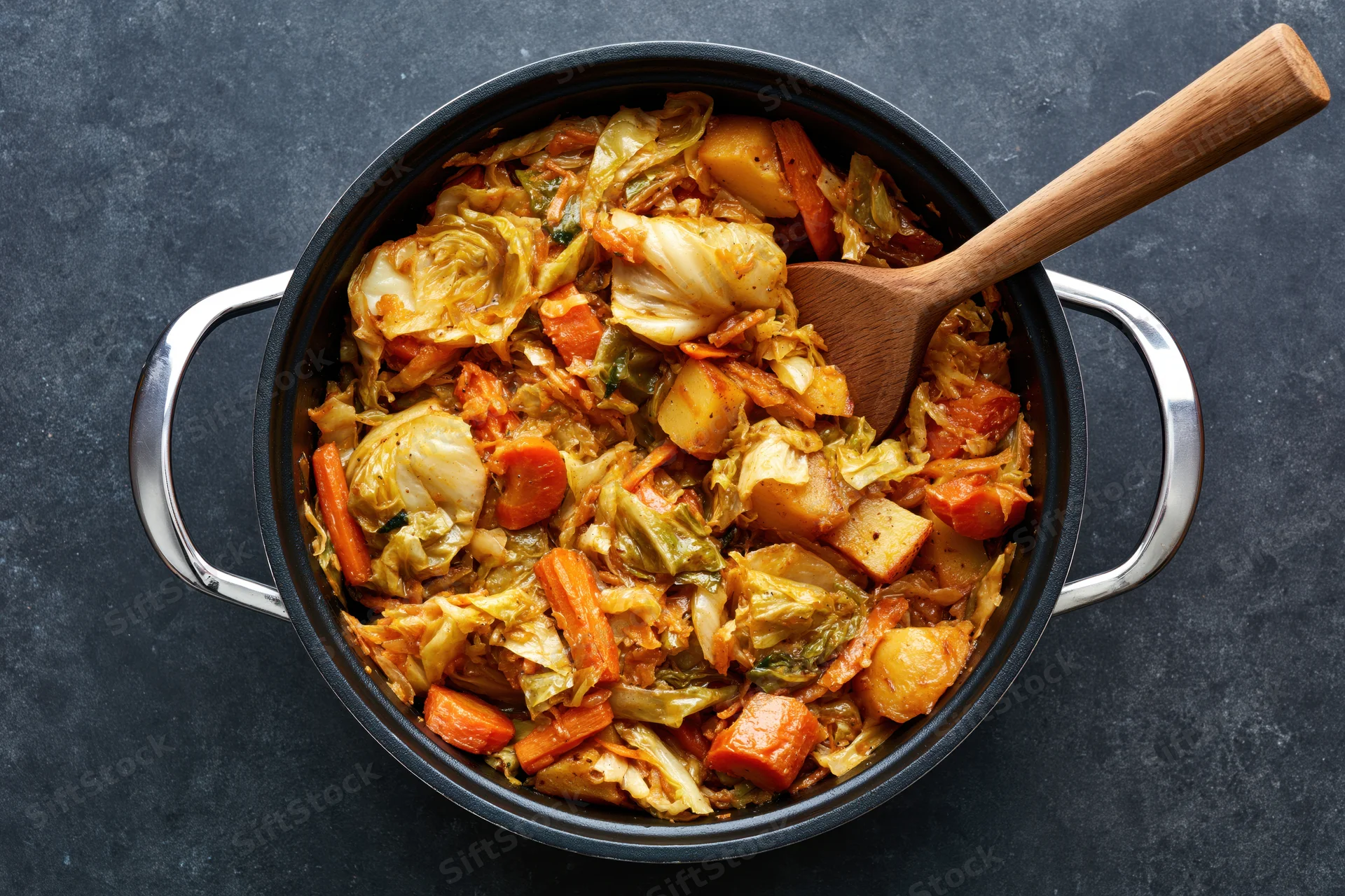 Overhead view of a black cast iron pot filled with sautéed cabbage, carrots and potatoes, with a wooden spoon resting inside, on a dark surface