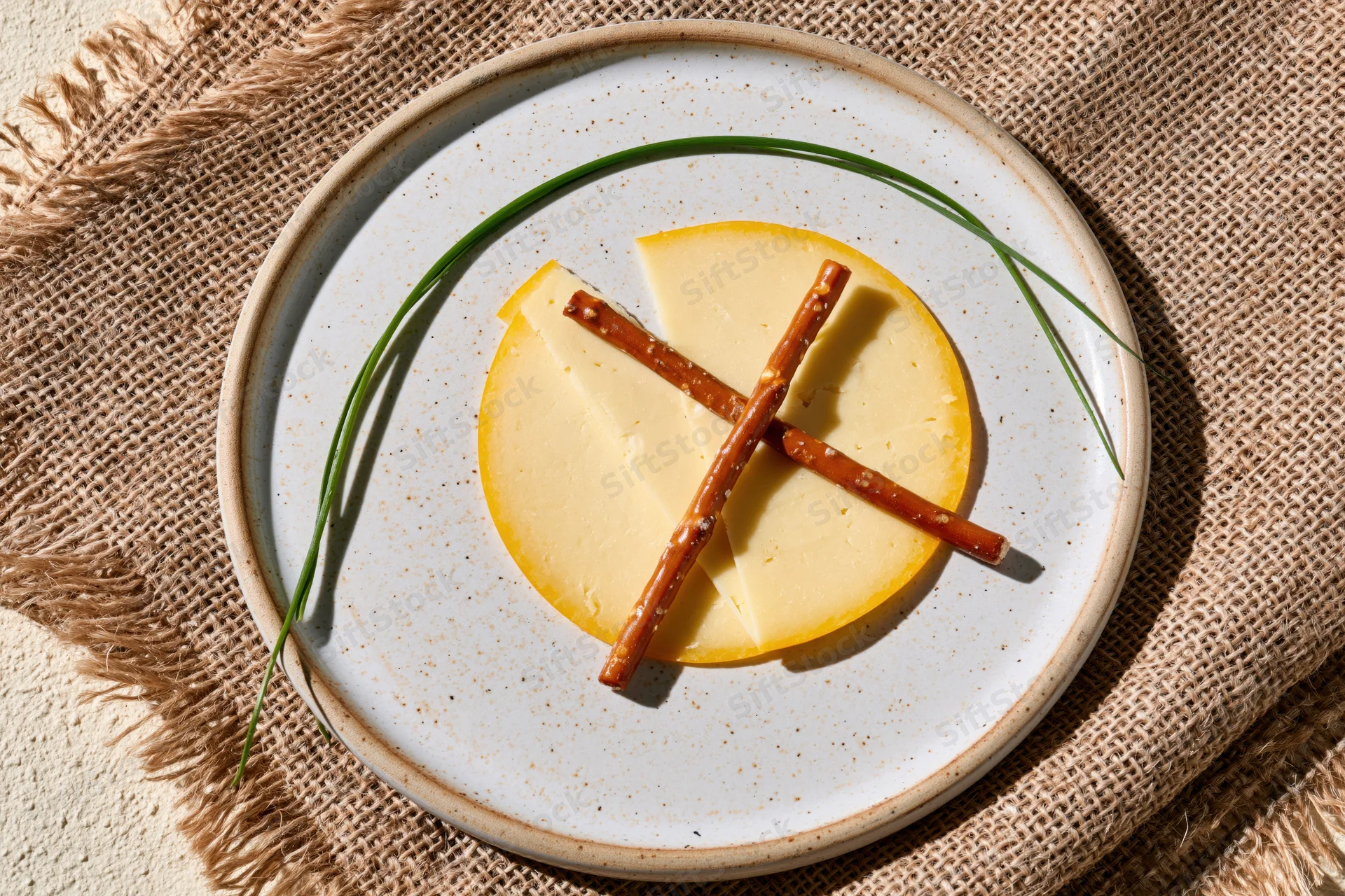 Round white speckled ceramic plate with cheese slices, crossed pretzels and chives on burlap textile
