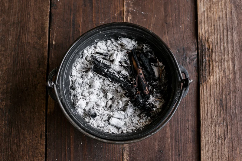Blackened cast iron pot filled with ash and charred wood on a dark wooden table