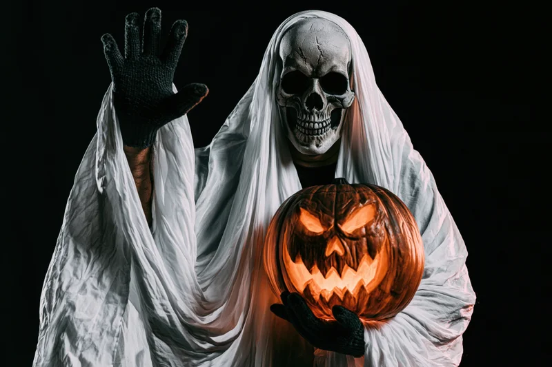 Ghost in white sheet costume with skull mask waving and holding carved pumpkin against black background