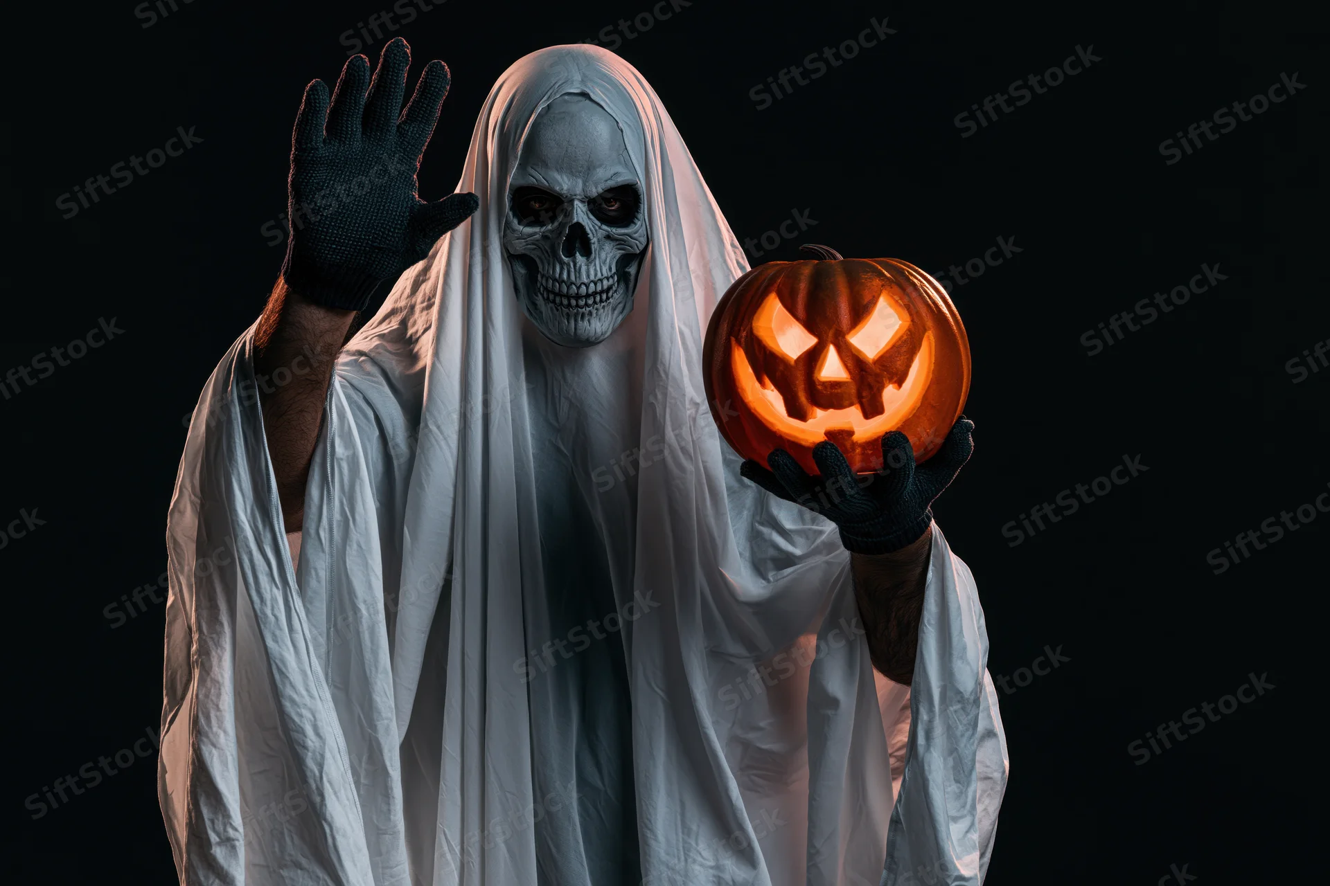 Ghost in white sheet costume with skull mask waving and holding glowing jack-o-lantern against dark background