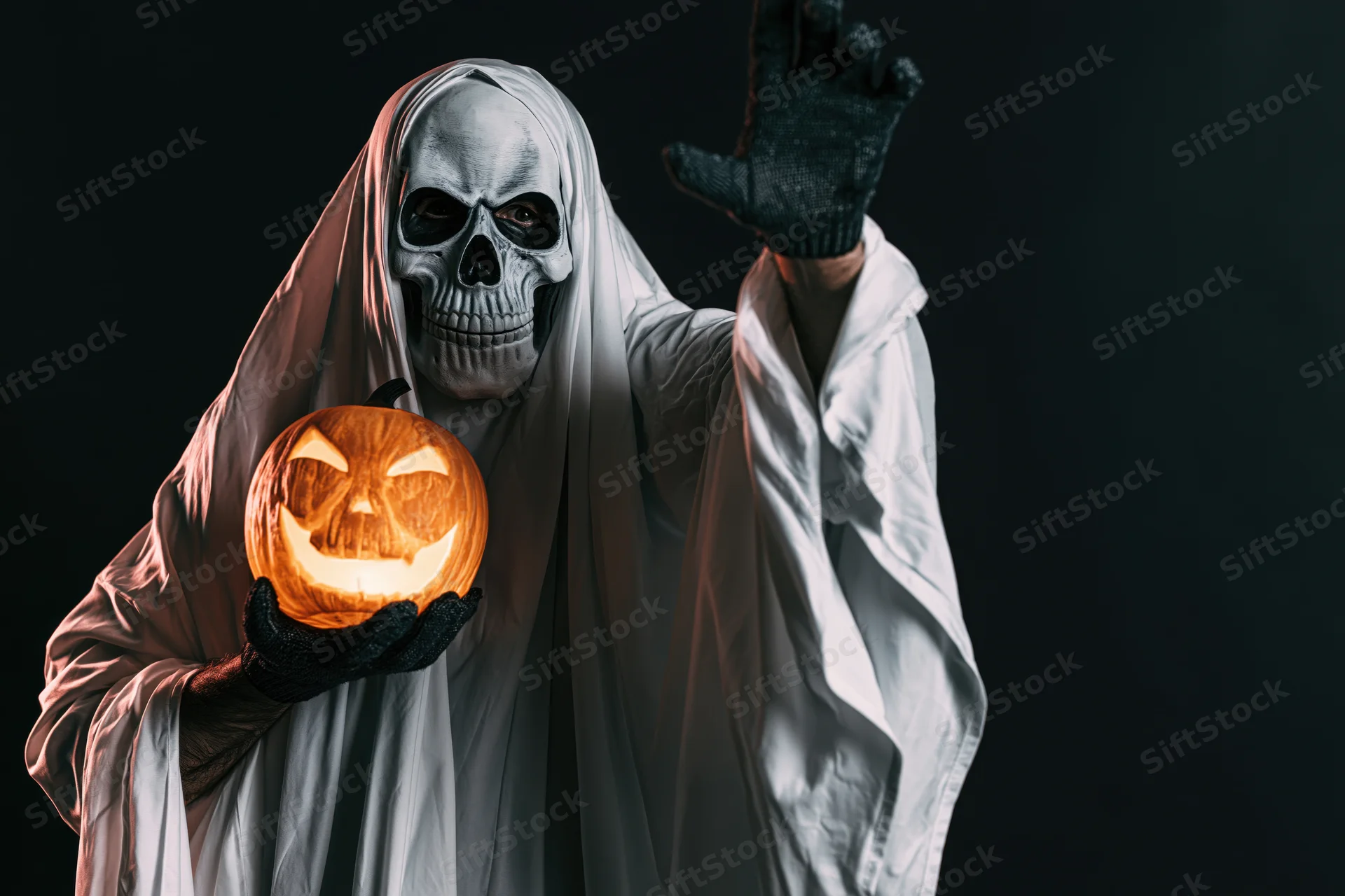 Hooded ghost in white sheet costume holding glowing jack-o-lantern and raising one hand in dark studio