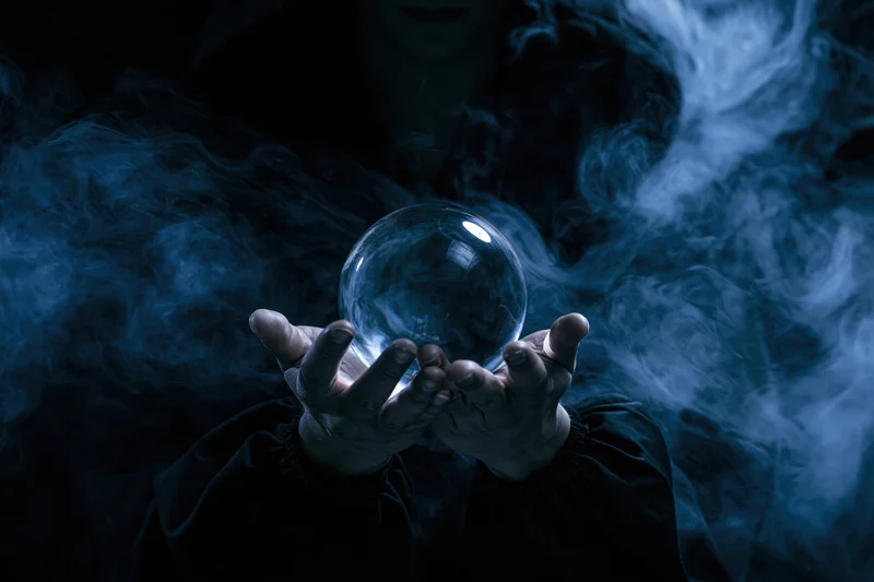 Woman holding a crystal ball in dark smoke, hooded and cloaked, hands cupped around the sphere