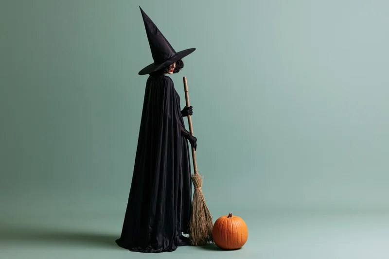 Woman in black witch costume holding broom beside pumpkin in studio