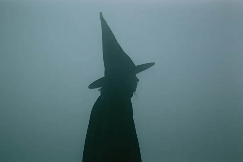 Silhouette of a witch in profile wearing a tall pointed hat and dark cloak in fog