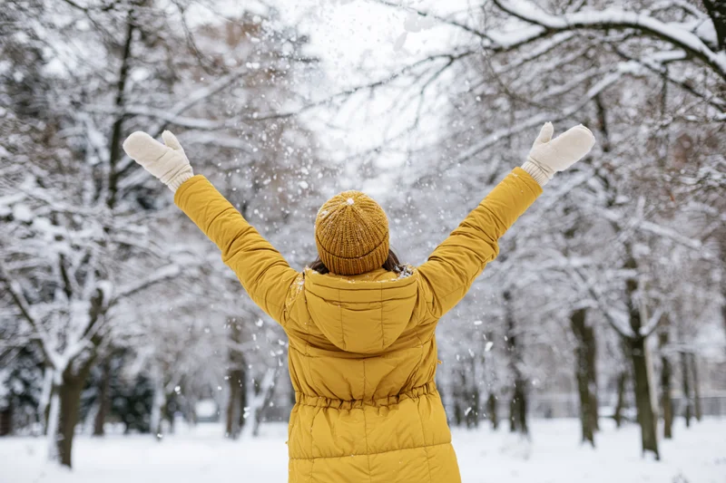 Woman in yellow puffer coat with arms raised tossing snow in snowy park