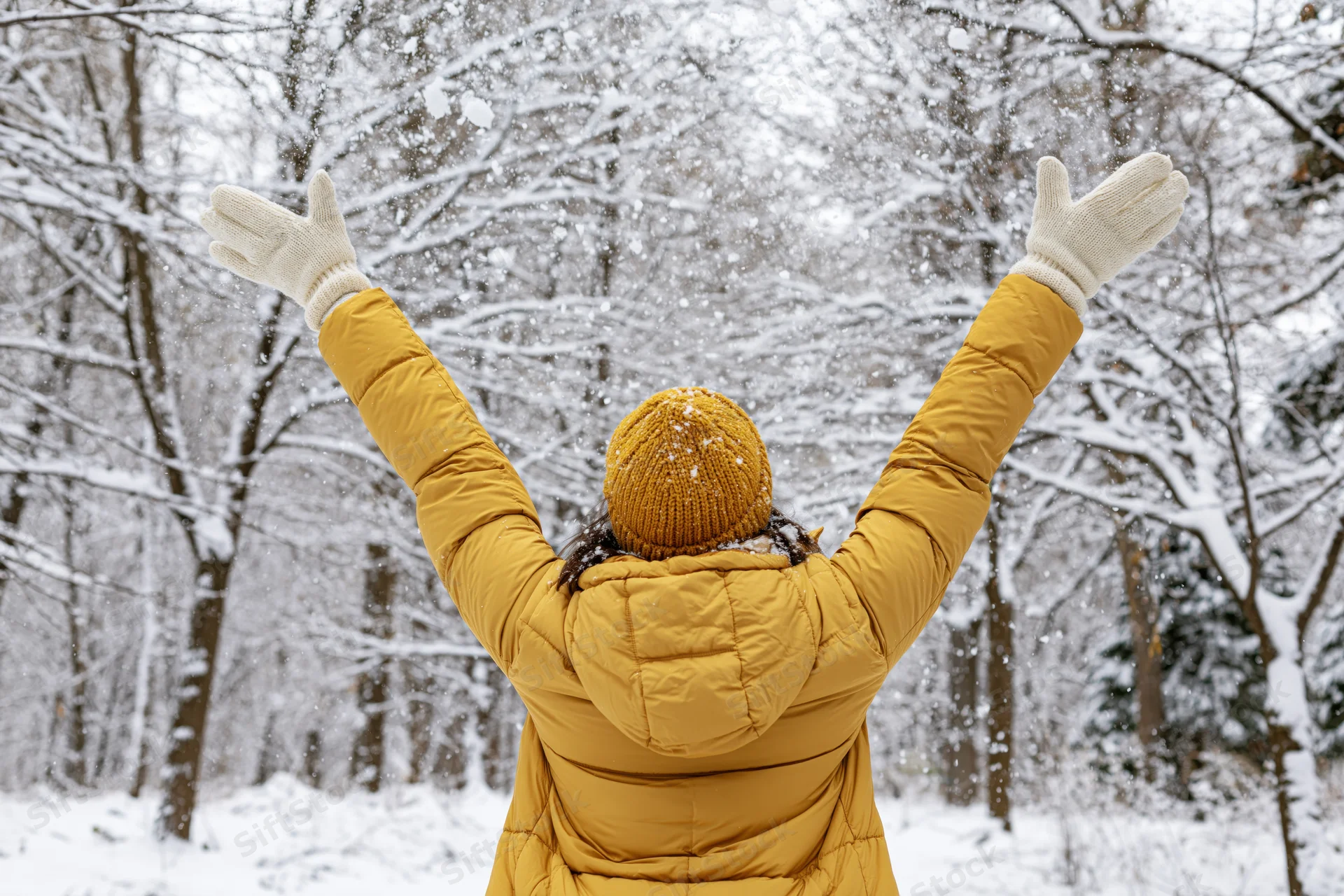 Woman in yellow puffer coat with arms raised tossing snow in snowy forest