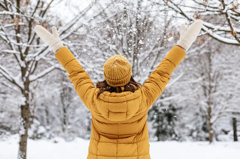 Woman in yellow puffer coat tossing snow in winter park with arms raised