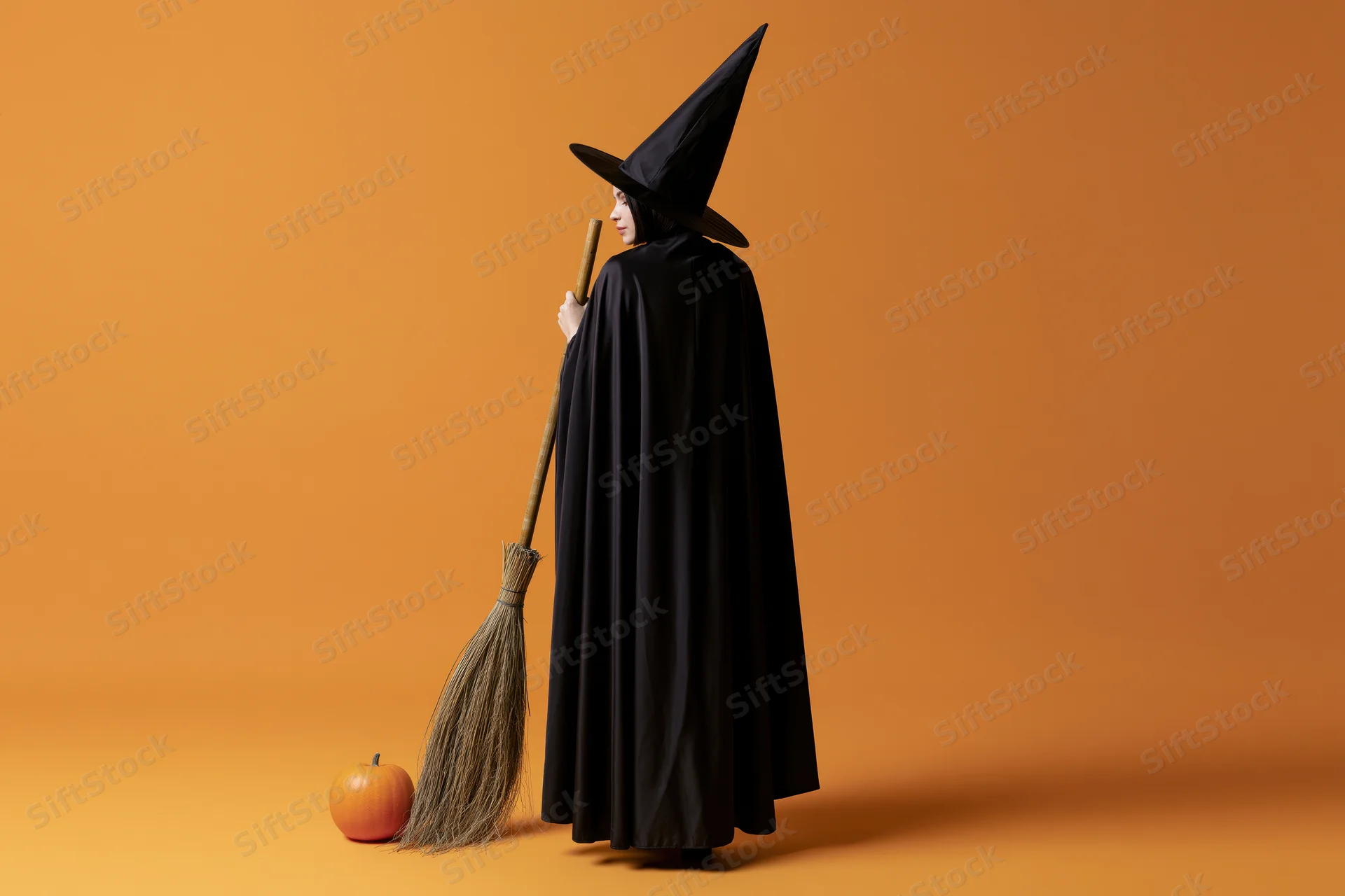 Woman in black witch costume holding broom beside pumpkin on orange background