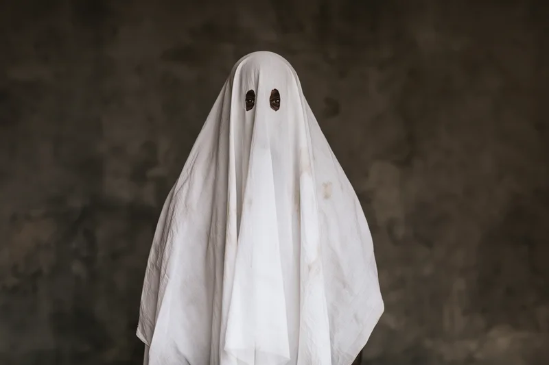 Person in worn white sheet ghost costume with eye cutouts standing against dark background