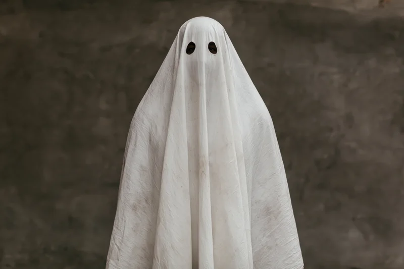 Person in worn white sheet ghost costume with eye cutouts standing against dark textured wall