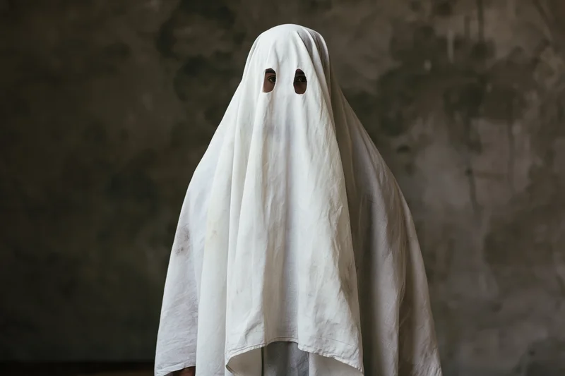 Person in worn white sheet ghost costume with eye holes standing against dark textured wall