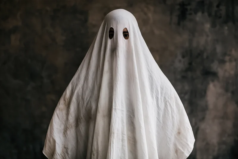 Person in worn white sheet ghost costume with eye holes against dark background