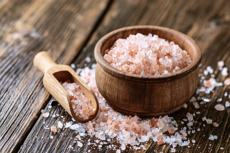 Pink himalayan salt crystals in a wooden bowl with a spoon on a rustic wooden surface