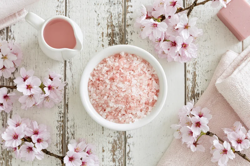 Pink bath salt crystals in a white ceramic bowl surrounded by cherry blossoms and towels on a wooden surface