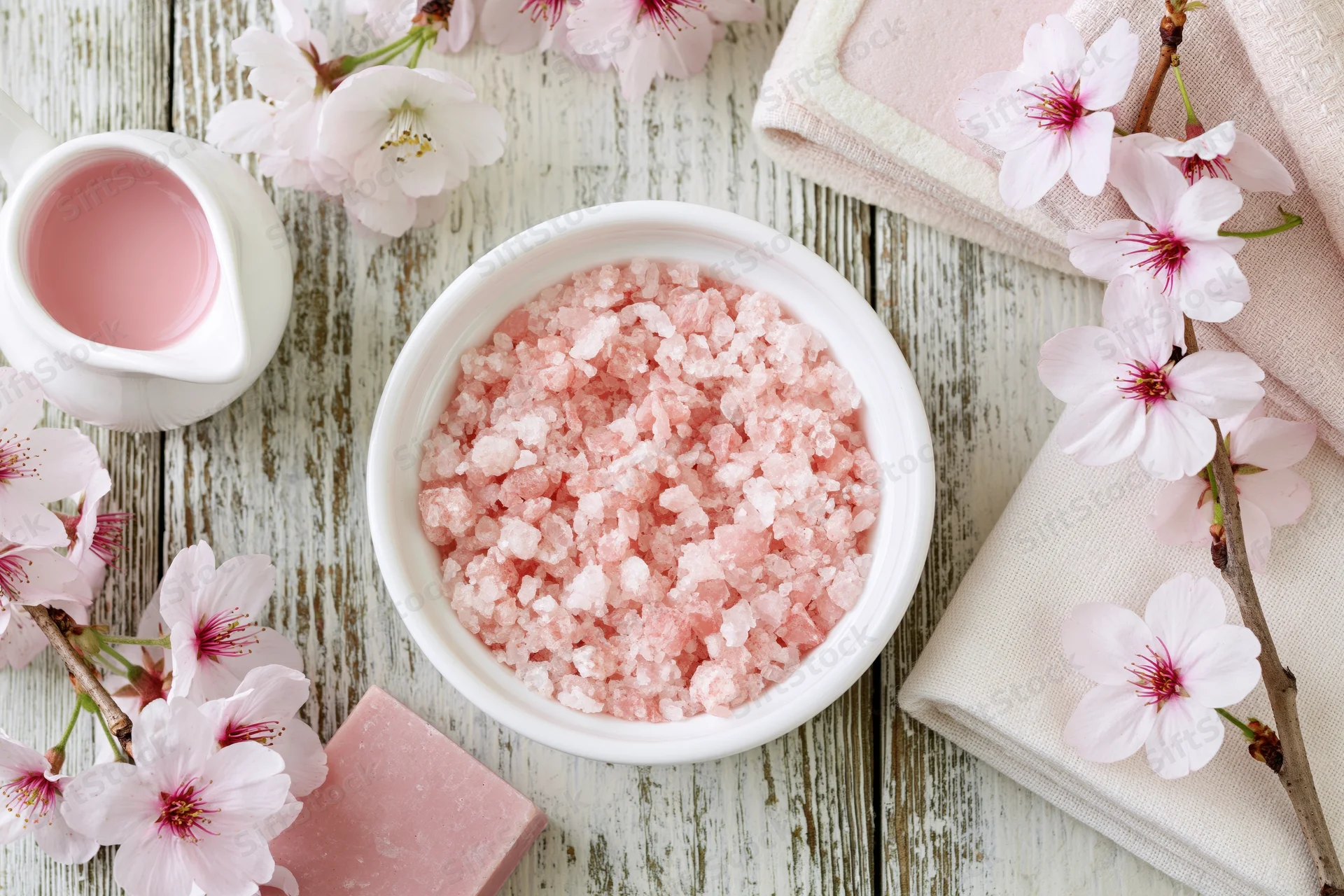 Pink bath salt crystals in a white ceramic bowl surrounded by cherry blossoms and towels on a wooden surface