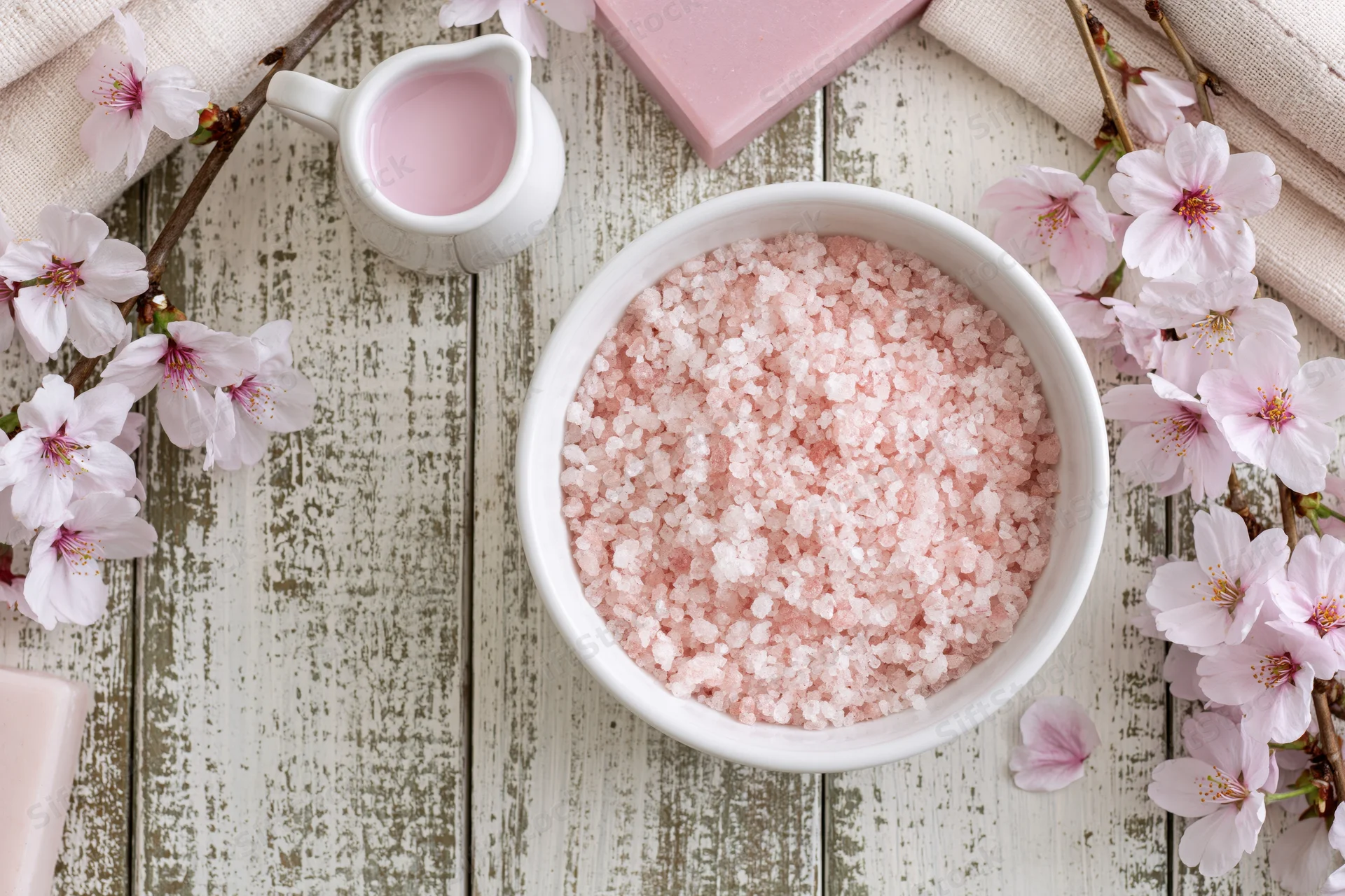 Pink bath salt crystals in a white ceramic bowl surrounded by cherry blossoms on a rustic wooden tabletop