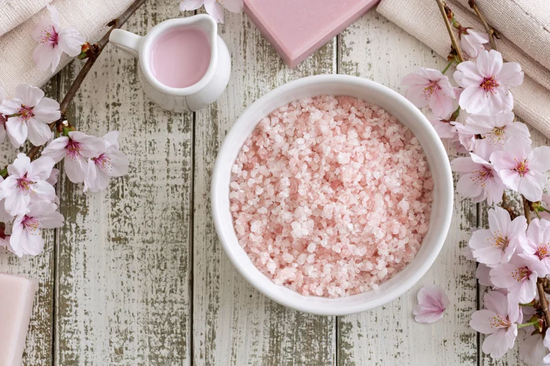 Pink bath salt crystals in a white ceramic bowl surrounded by cherry blossoms on a rustic wooden tabletop