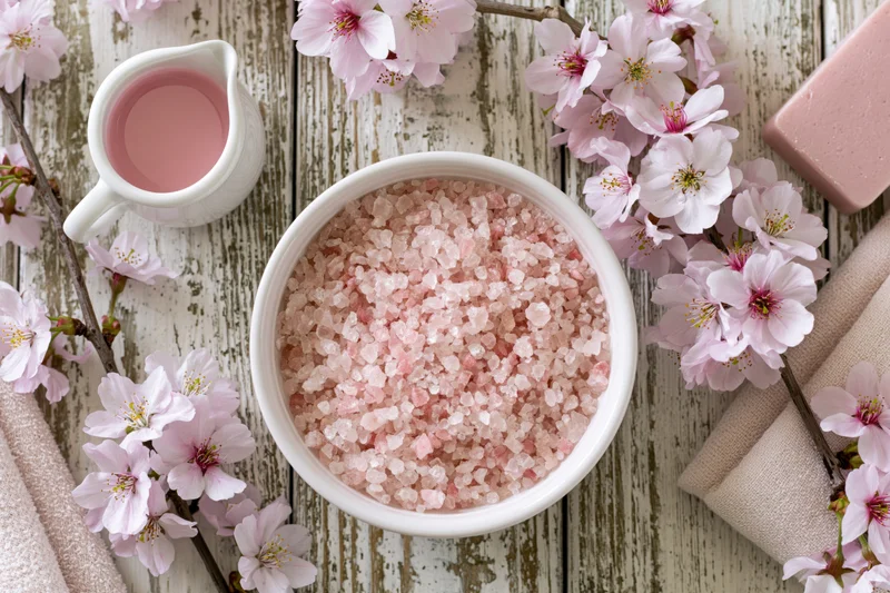 Pink bath salt crystals in a white ceramic bowl surrounded by cherry blossoms on a rustic wooden table