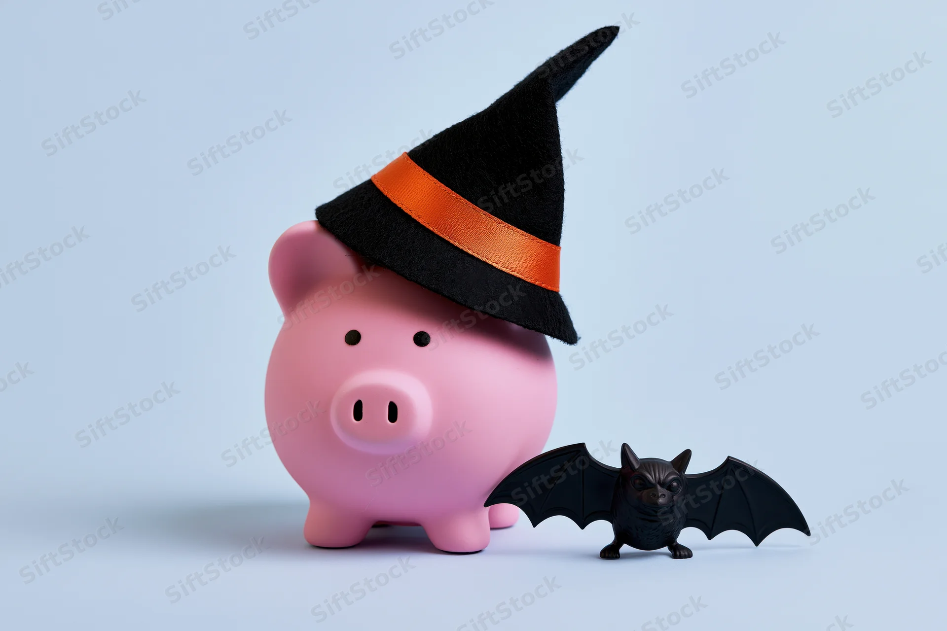 Pink ceramic piggy bank wearing a black witch hat with orange band beside a small black bat figurine on light blue background