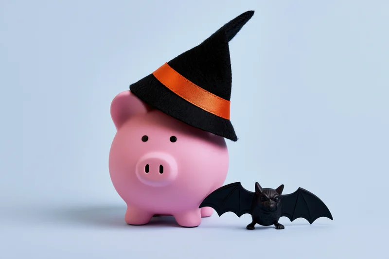 Pink ceramic piggy bank wearing a black witch hat with orange band beside a small black bat figurine on light blue background