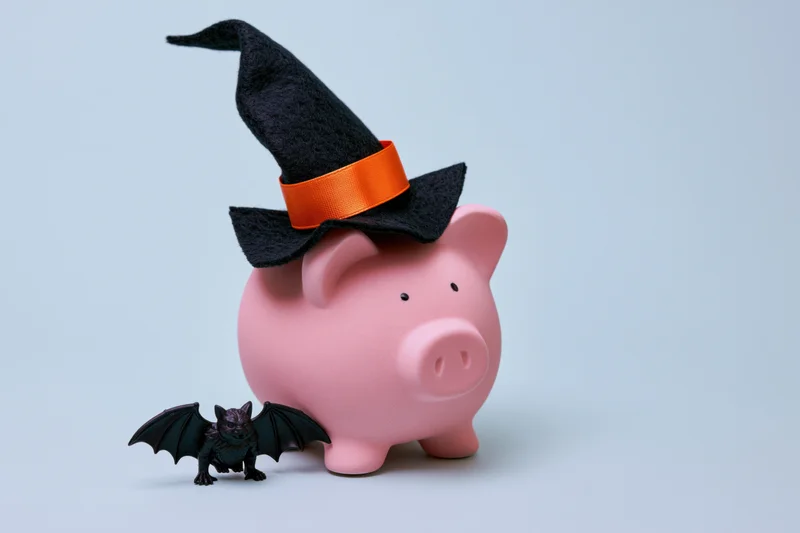 Pink ceramic piggy bank wearing a black witch hat with orange band, accompanied by a small black bat toy, on a plain light background