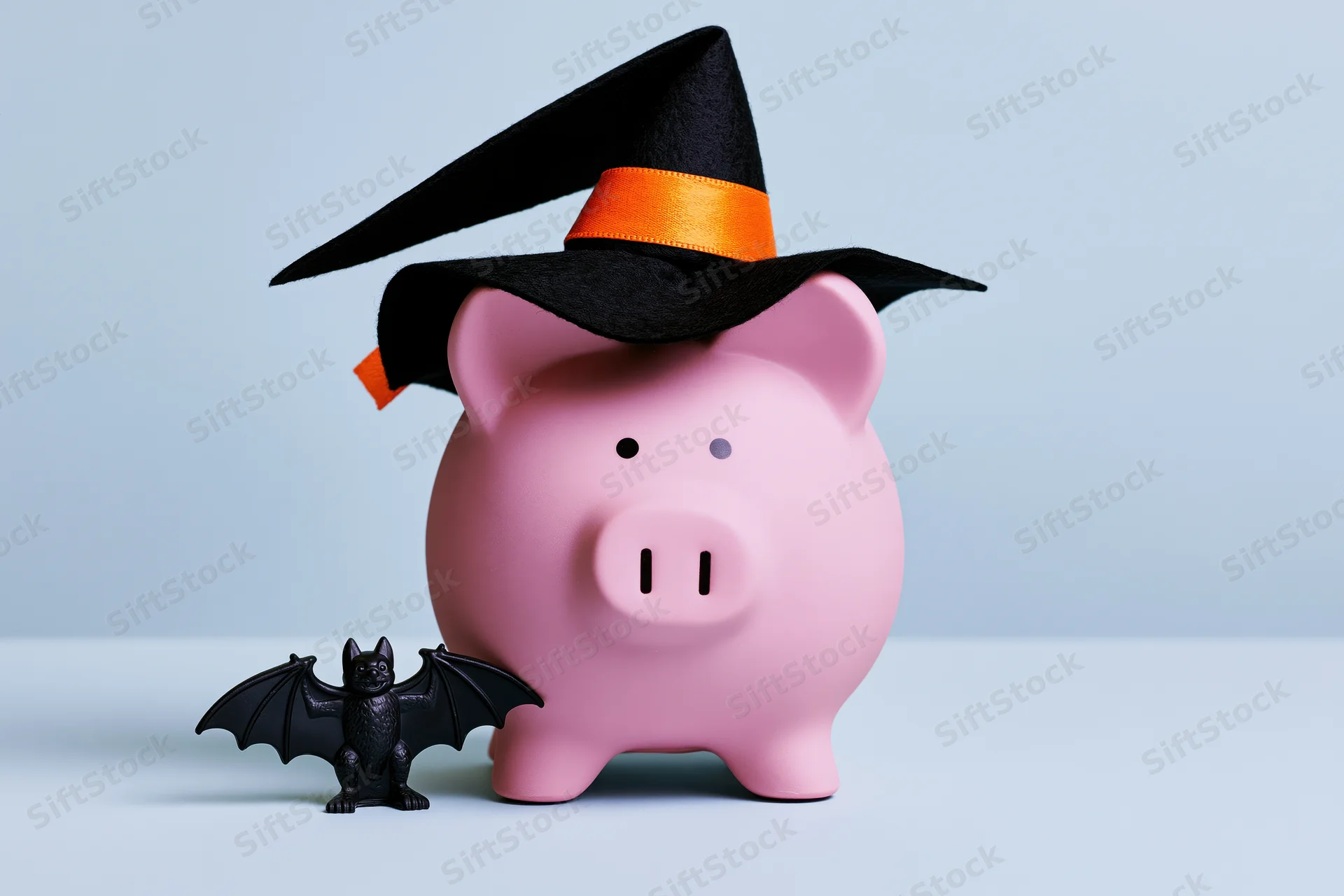 Pink piggy bank wearing witch hat with small bat figurine beside it on light blue surface
