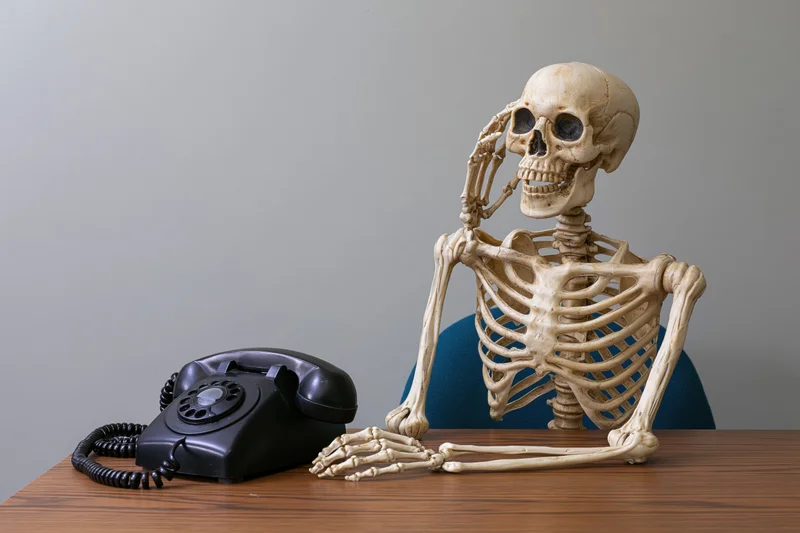Human skeleton prop sitting at desk holding telephone receiver near vintage rotary phone