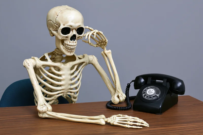 Skeleton sitting at a desk with a vintage rotary telephone, hand to ear as if listening