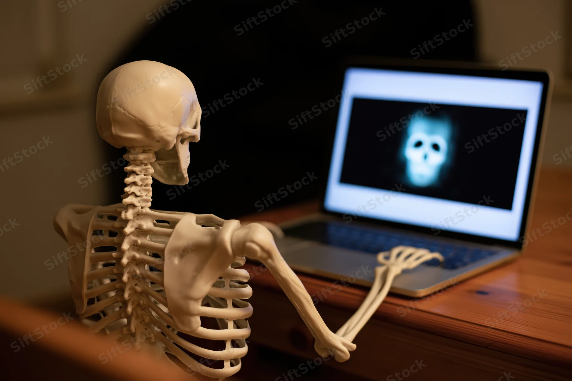 Plastic anatomy skeleton model sitting at wooden desk using laptop with glowing skull on screen