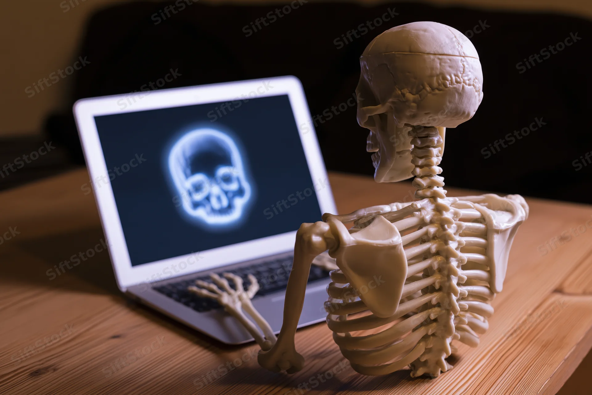 Plastic skeleton model sitting at wooden desk using laptop with glowing skull on screen