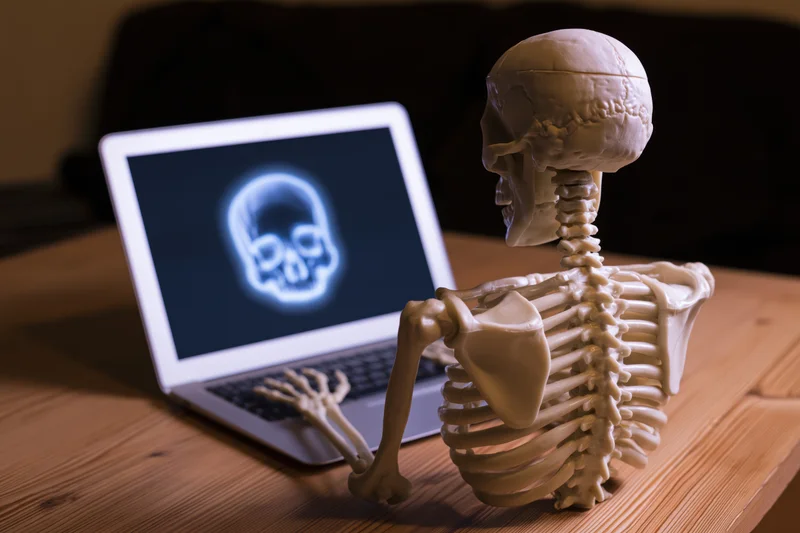 Plastic skeleton model sitting at wooden desk using laptop with glowing skull on screen