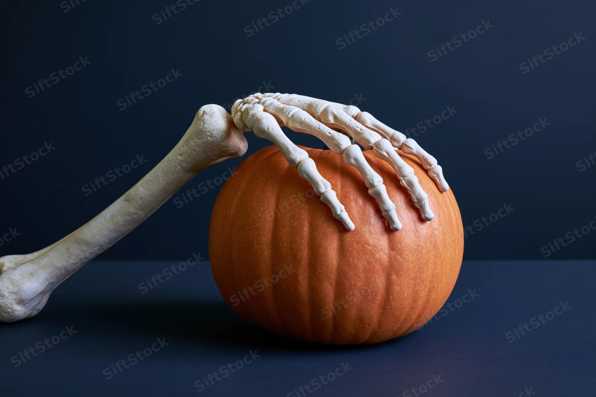 Plastic skeleton hand gripping a small orange pumpkin on a dark blue surface