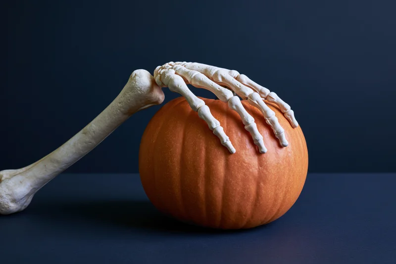 Plastic skeleton hand gripping a small orange pumpkin on a dark blue surface