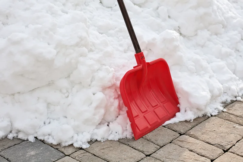Red plastic snow shovel resting in snow on a brick sidewalk