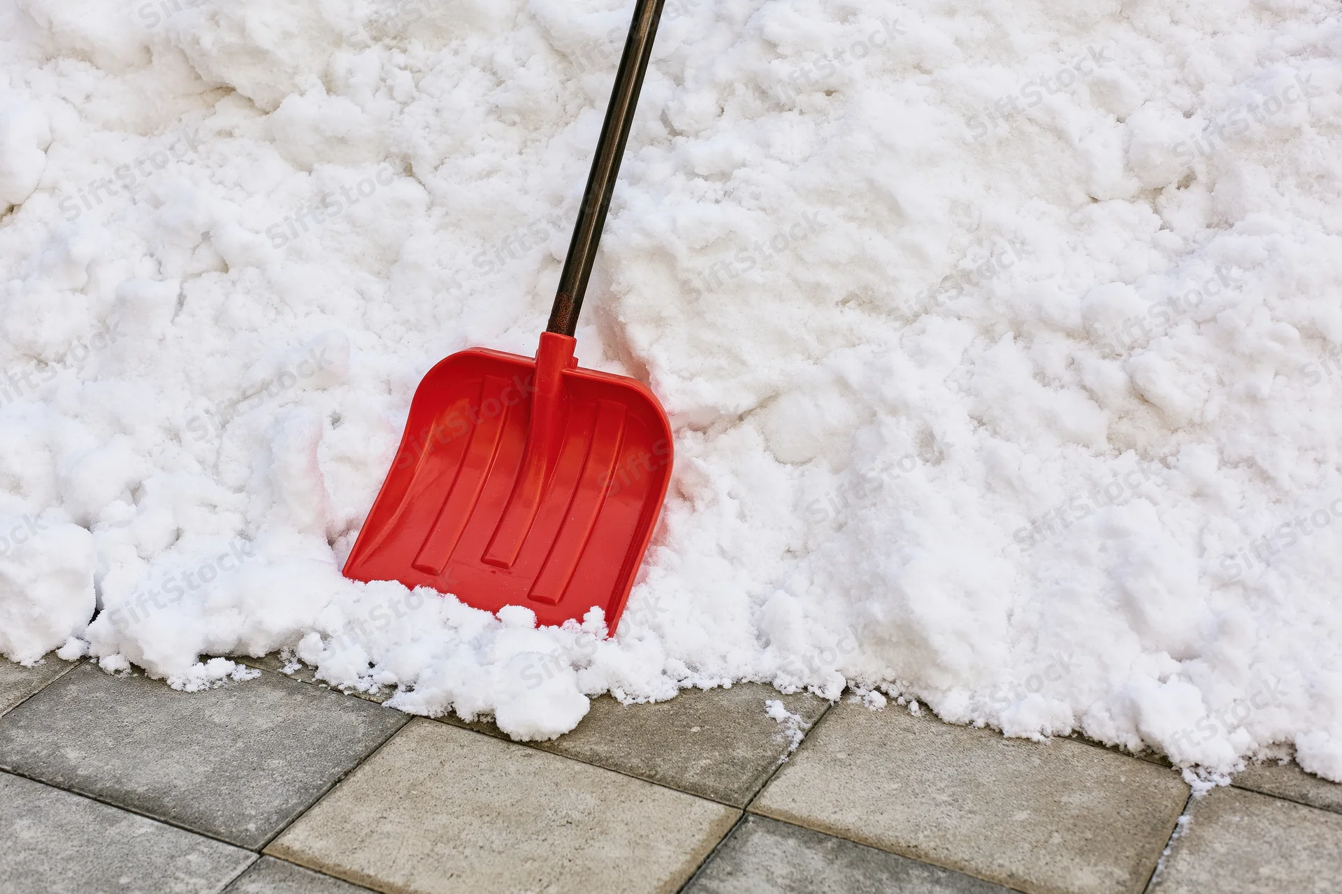 Red plastic snow shovel with dark metal handle resting in snow on tiled pavement