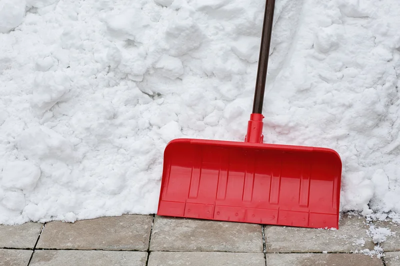 Red plastic snow shovel with dark metal handle resting against a snowbank on paved ground