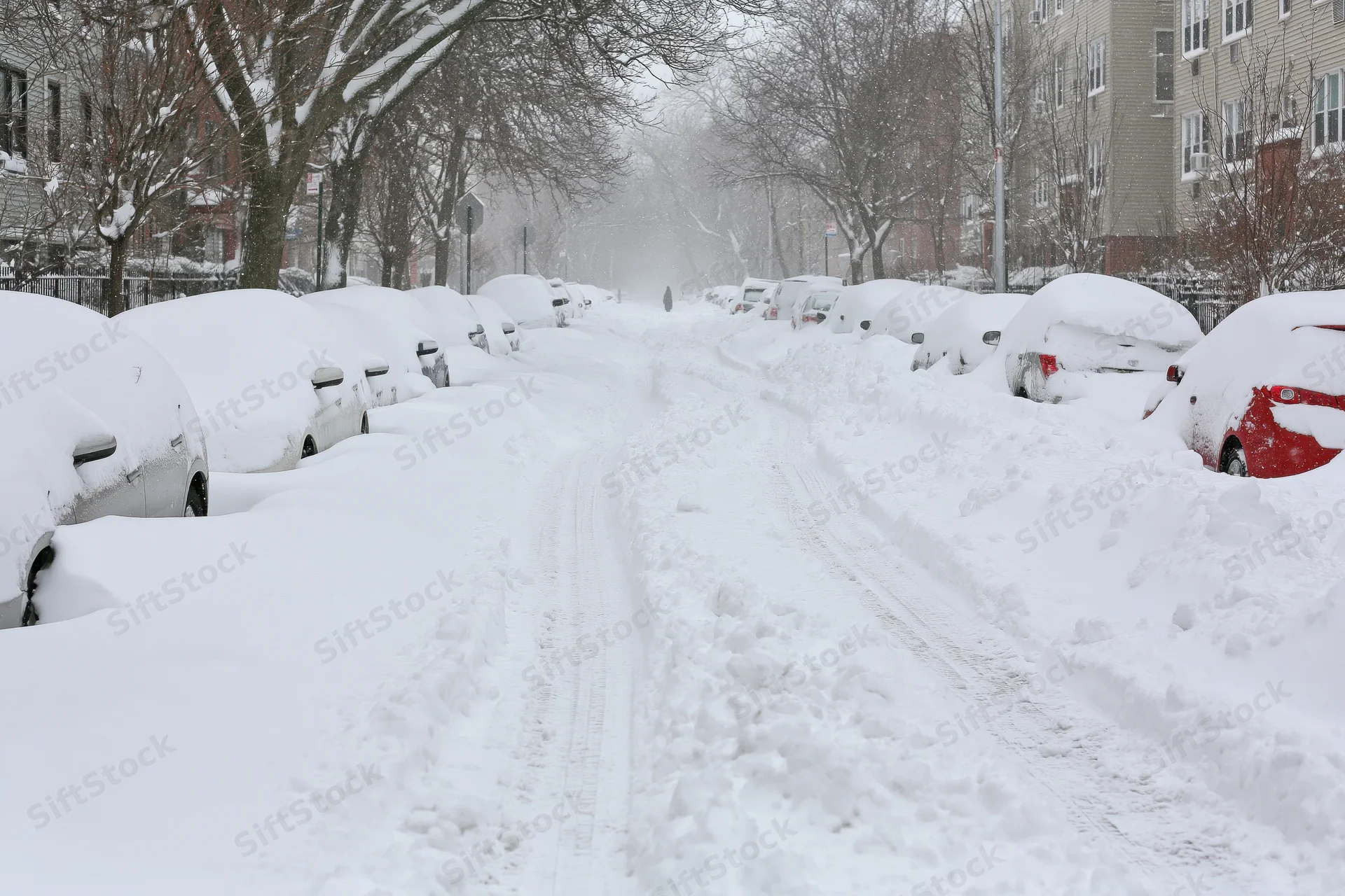Snow-covered residential street with parked cars and tire tracks during heavy snowstorm