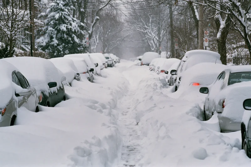 Snow-covered residential street with parked cars and footprints in the snow during heavy snowstorm