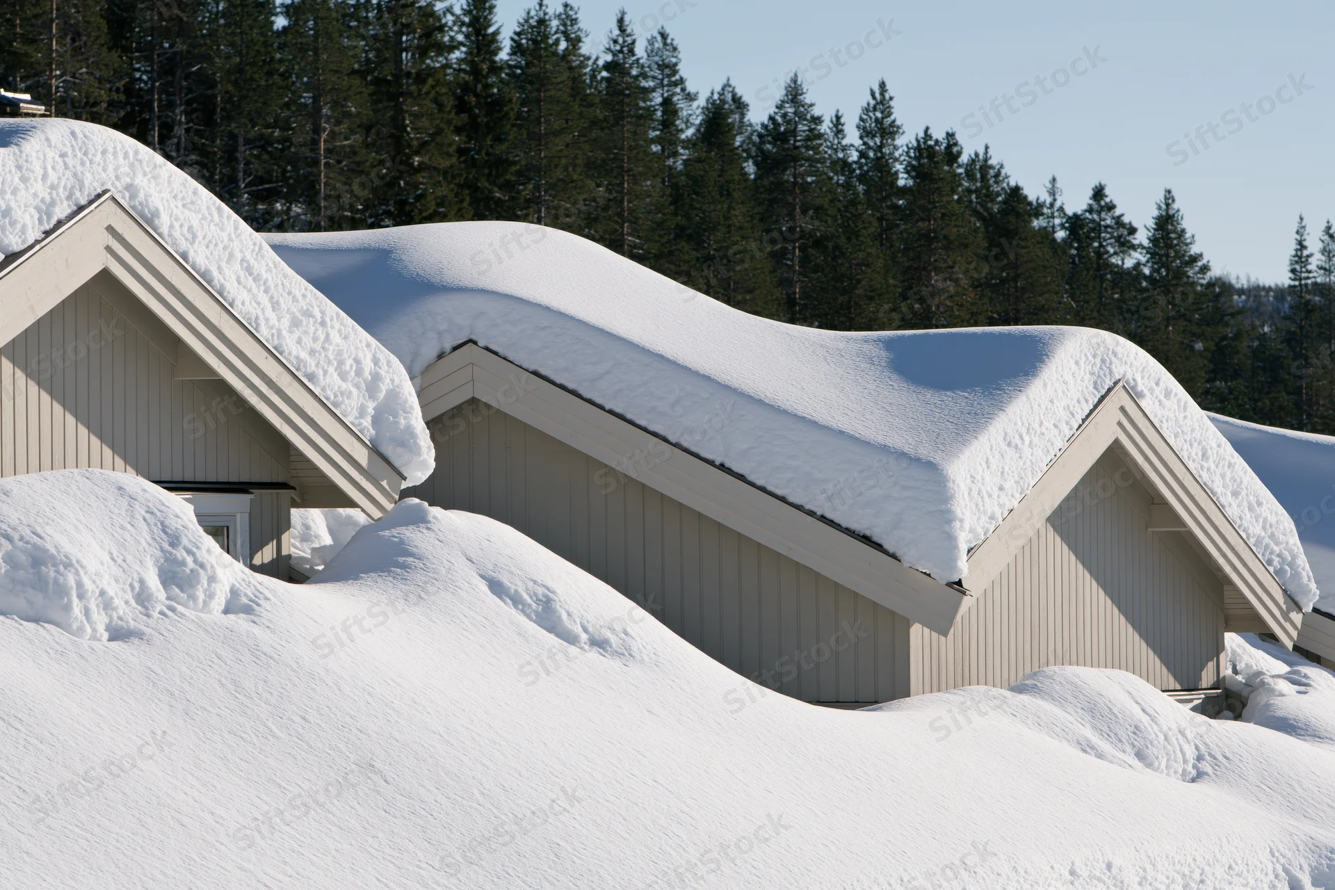 Row of pale grey scandinavian houses with thick snow on roofs and ground surrounded by pine forest
