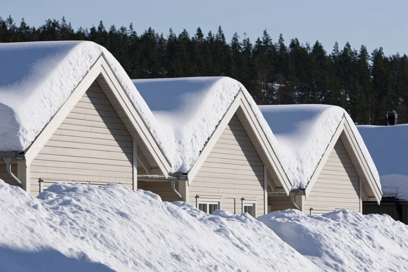 Row of pale grey scandinavian houses with snow-covered roofs and snowdrifts in front, set against a forested background under a clear sky