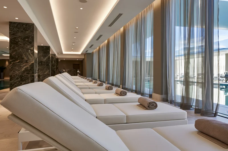 Row of white padded loungers with rolled towels arranged in a modern spa interior near large windows overlooking a pool