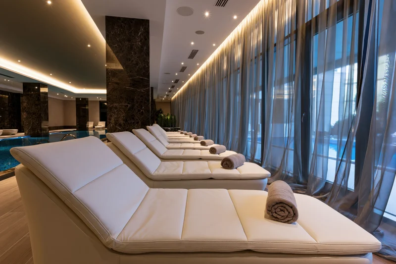 Row of white padded loungers with rolled towels arranged by a poolside window in a luxury spa interior
