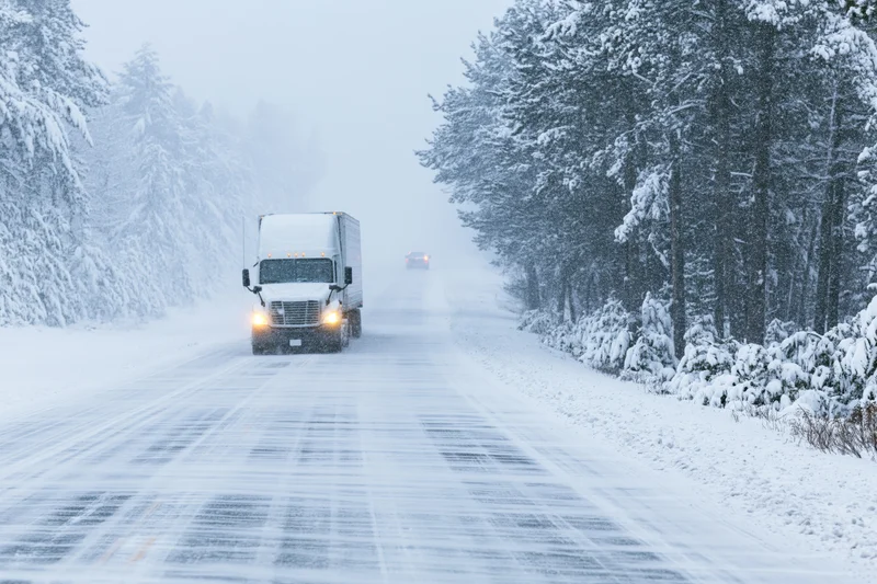 Semi-truck driving toward camera on snow-covered two-lane highway flanked by snow-laden trees
