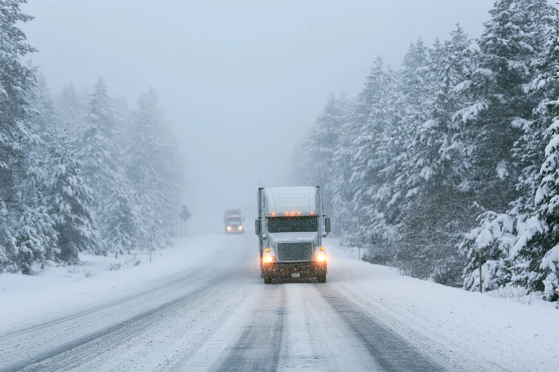 Semi-truck driving toward camera on snow-covered two-lane highway flanked by snow-laden pine trees