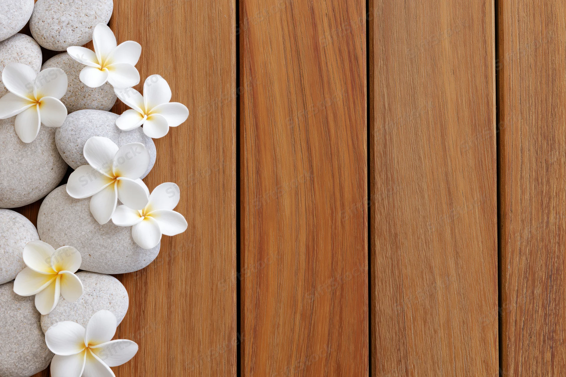 Smooth stones and white plumeria flowers arranged along the left edge of a wooden deck, leaving copyspace on the right
