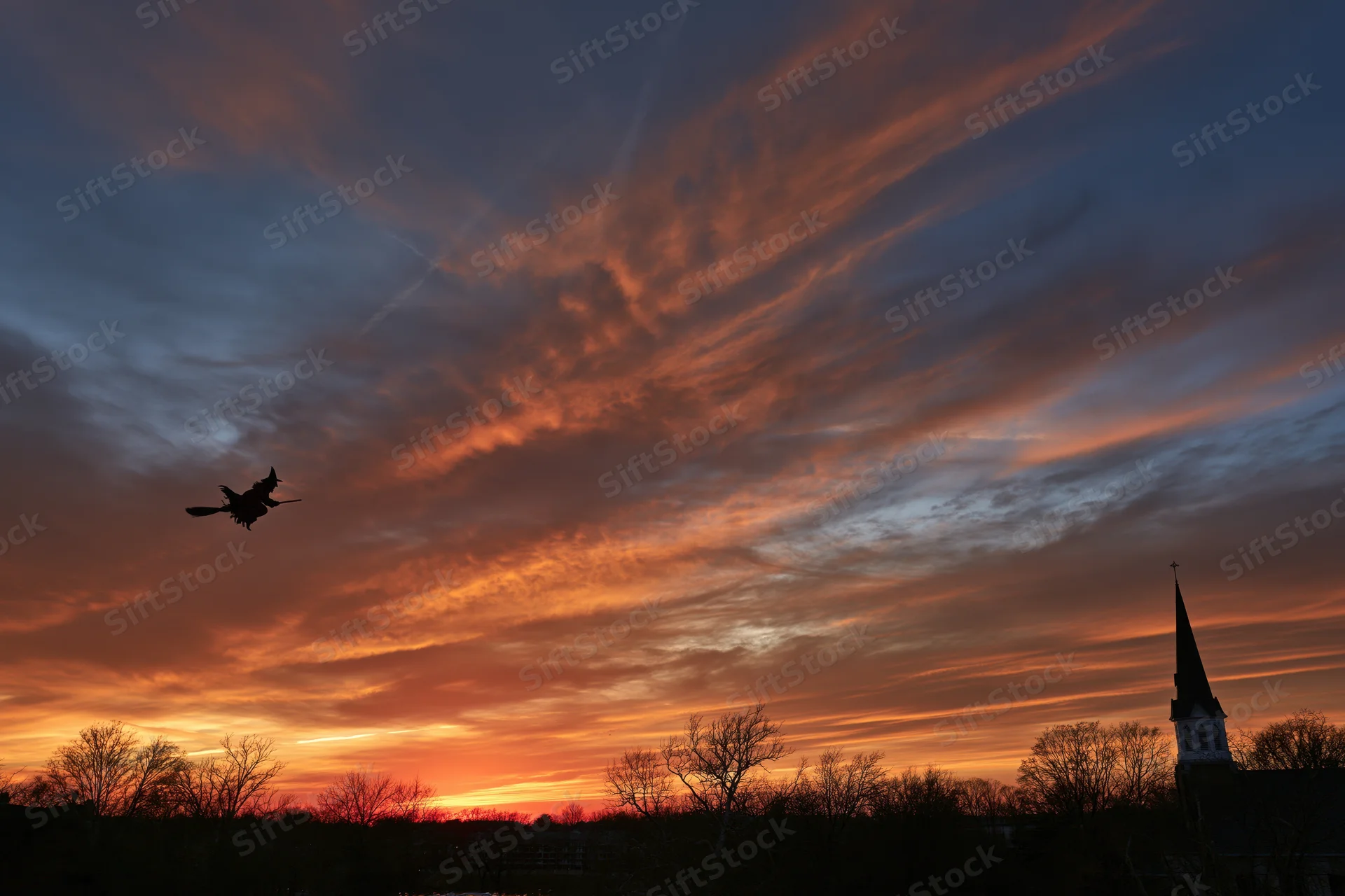 Witch silhouette flying on broomstick across sunset sky over rural landscape with church steeple
