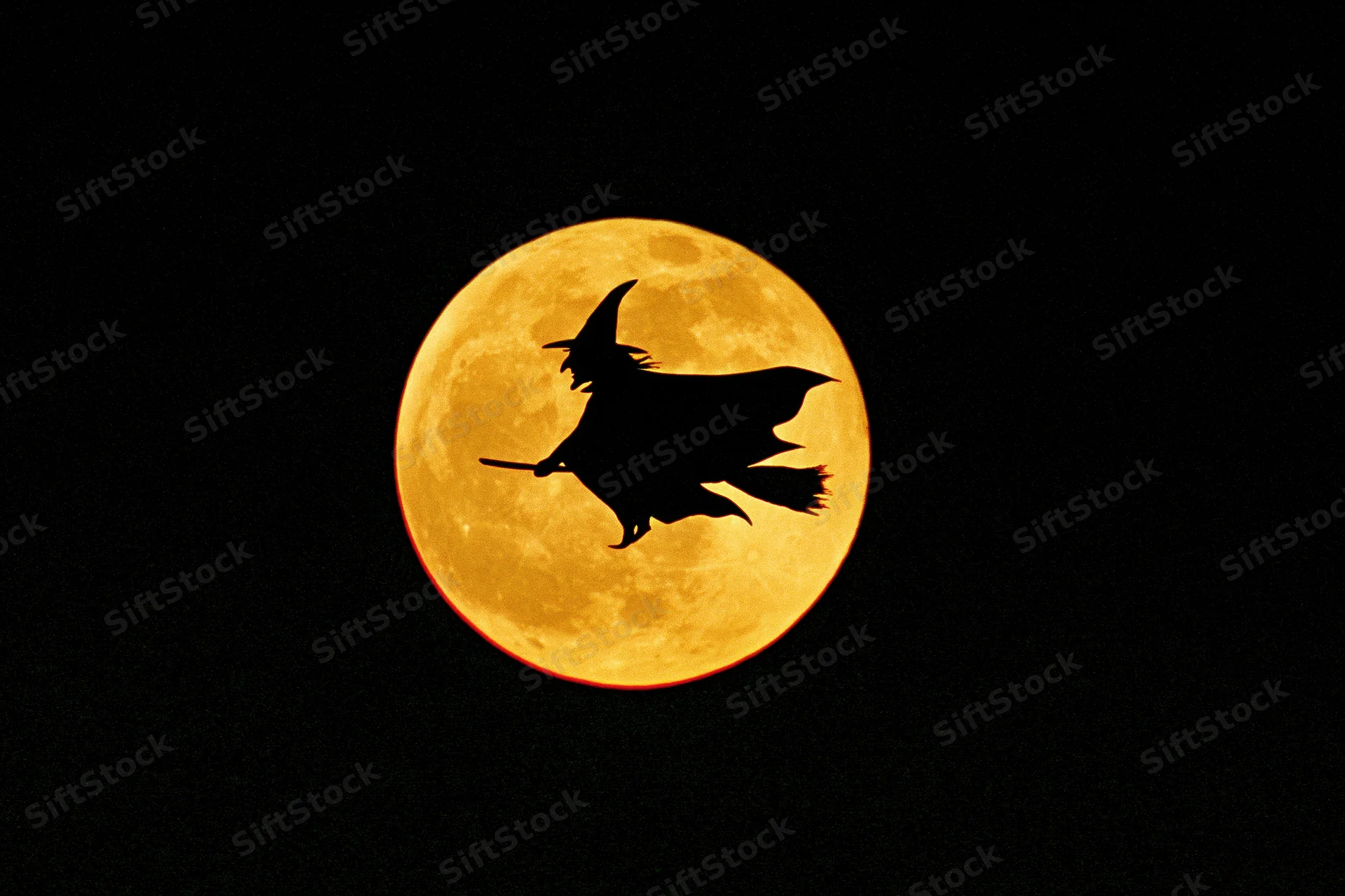 Silhouette of a witch riding a broomstick against a full orange moon at night