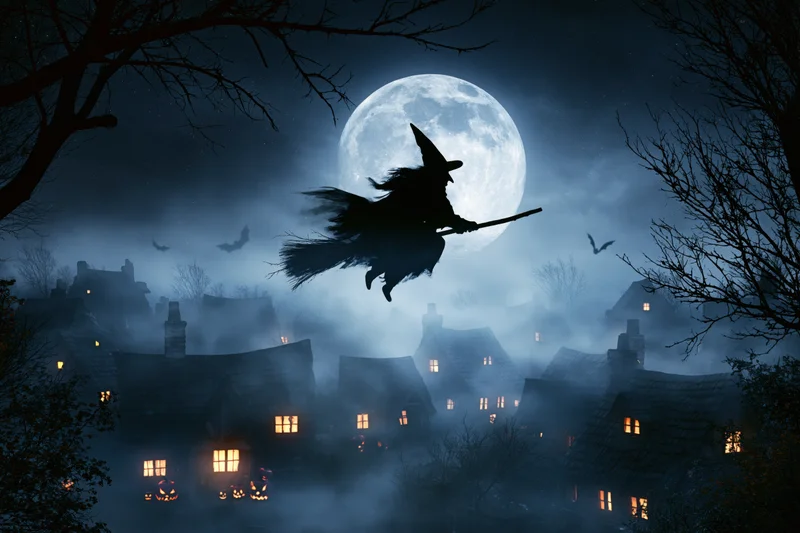 Witch flying on broomstick over foggy village at night with full moon and jack-o'-lanterns lit in windows
